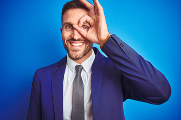 Close up picture of young handsome business man over blue isolated background doing ok gesture with hand smiling, eye looking through fingers with happy face.