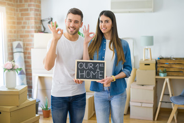 Fototapeta na wymiar Young beautiful couple holding blackboard with our first home text at new house doing ok sign with fingers, excellent symbol