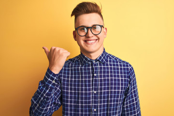 Young handsome man wearing casual shirt and glasses over isolated yellow background smiling with happy face looking and pointing to the side with thumb up.