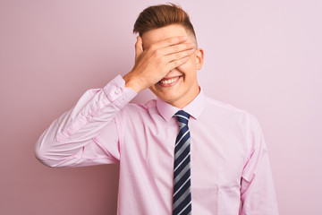 Young handsome businessman wearing shirt and tie standing over isolated pink background smiling and laughing with hand on face covering eyes for surprise. Blind concept.