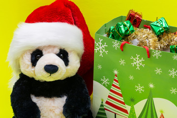 Bag with Gifts and Panda Bear plush toy wearing Red Christmas Santa Hat on bright yellow background. Christmas, New Year, Winter Holidays concept.