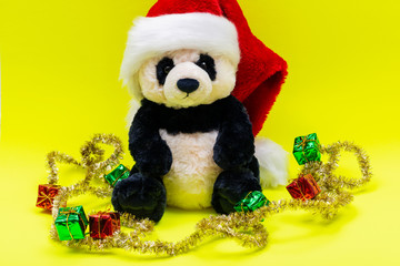 Panda Bear plush toy with Red Christmas Santa Hat surrounded by Tinsel Garlands on bright yellow background. Winter Holidays concept.