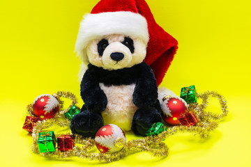 Panda Bear plush toy with Red Christmas Santa Hat surrounded by Tinsel Garland and Vintage Ornaments on bright yellow background. Holidays concept.