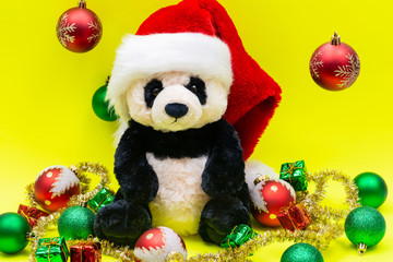 Panda Bear plush toy with Red Christmas Santa Hat surrounded by Tinsel Garland and Vintage Ornaments on bright yellow background. Holidays concept.