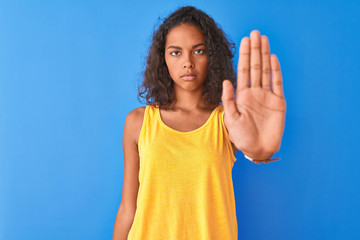 Young brazilian woman wearing yellow t-shirt standing over isolated blue background doing stop sing...