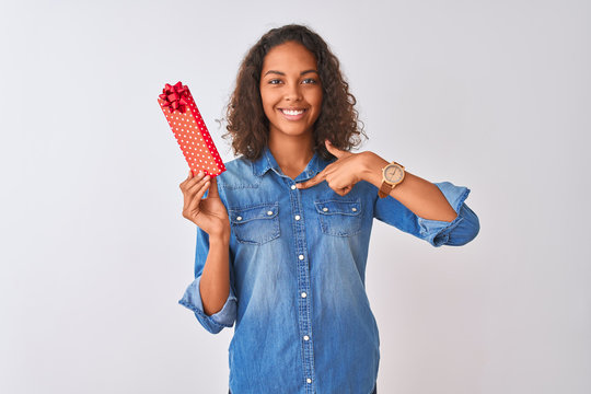 Young brazilian woman holding valentine gift standing over isolated white background with surprise face pointing finger to himself