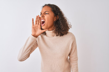 Young brazilian woman wearing turtleneck sweater standing over isolated white background shouting and screaming loud to side with hand on mouth. Communication concept.