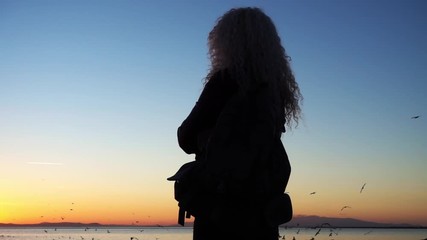 Woman standing on the beach and looking at birds flying at sunset.