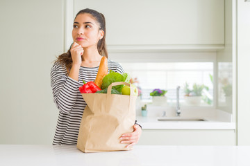 Young woman holding paper bag full of groceries serious face thinking about question, very confused...