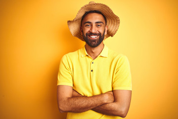 Young indian man on vacation wearing summer hat standing over isolated yellow background happy face smiling with crossed arms looking at the camera. Positive person.