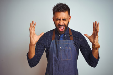 Fototapeta premium Young indian employee man wearing apron uniform standing over isolated white background celebrating mad and crazy for success with arms raised and closed eyes screaming excited. Winner concept