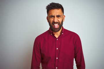Young indian man wearing red elegant shirt standing over isolated grey background sticking tongue out happy with funny expression. Emotion concept.