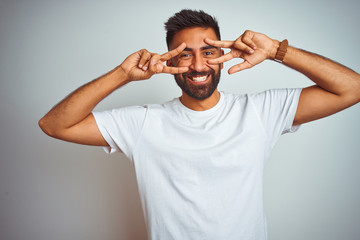 Young indian man wearing t-shirt standing over isolated white background Doing peace symbol with...