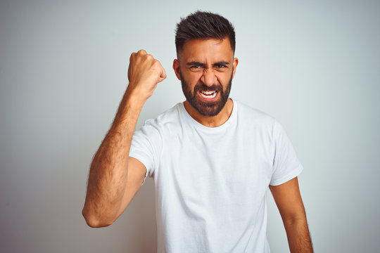 Young indian man wearing t-shirt standing over isolated white background angry and mad raising fist frustrated and furious while shouting with anger. Rage and aggressive concept.