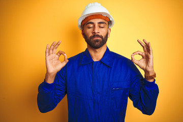 Handsome indian worker man wearing uniform and helmet over isolated yellow background relax and smiling with eyes closed doing meditation gesture with fingers. Yoga concept.