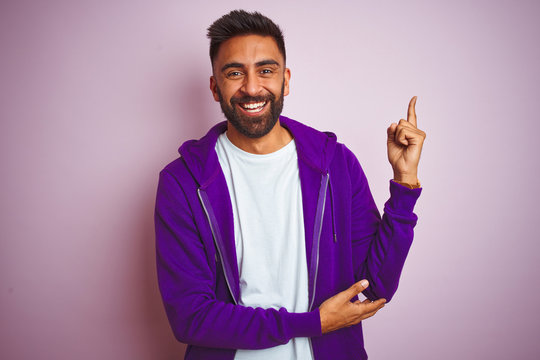 Young indian man wearing purple sweatshirt standing over isolated pink background with a big smile on face, pointing with hand and finger to the side looking at the camera.