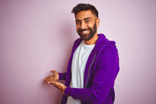 Young indian man wearing purple sweatshirt standing over isolated pink background Inviting to enter smiling natural with open hand