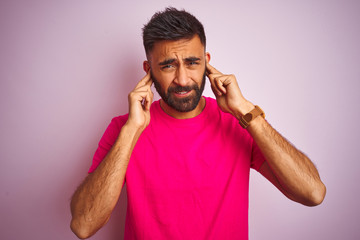 Fototapeta na wymiar Young indian man wearing t-shirt standing over isolated pink background covering ears with fingers with annoyed expression for the noise of loud music. Deaf concept.