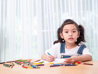 Asian little cute girl sitting and use color pencil drawing cartoon in white paper on wooden table. Preschool lovely kid with  drawing at paper near the window. Learning and education of kid.