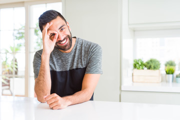 Handsome hispanic man wearing casual t-shirt at home doing ok gesture with hand smiling, eye looking through fingers with happy face.