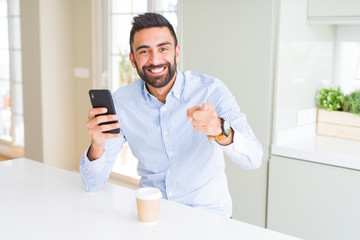 Handsome hispanic business man drinking coffee and using smartphone pointing with finger to the camera and to you, hand sign, positive and confident gesture from the front