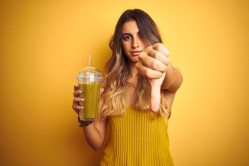 Young beautiful woman drinking detox green smoothy over yellow isolated background with angry face,...
