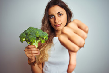 Young beautiful woman eating broccoli over grey isolated background with angry face, negative sign showing dislike with thumbs down, rejection concept
