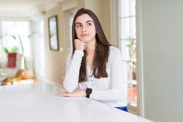 Beautiful young woman sitting on white table at home with hand on chin thinking about question, pensive expression. Smiling with thoughtful face. Doubt concept.