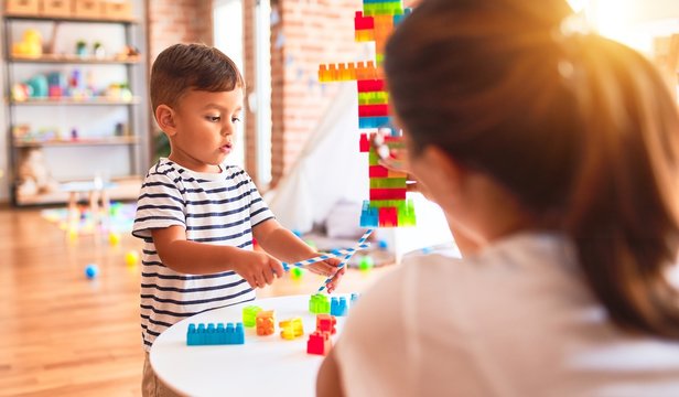 Beautiful teacher and toddler boy playing with construction blocks bulding tower at kindergarten