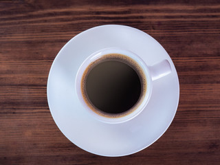White cup full of aromatic coffee, close up, top view. White coffee cup and saucer on brown table. Texture of wooden desk. Selective soft focus. Blurred background