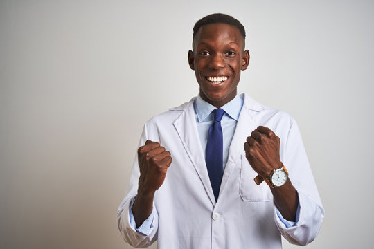 Young african american doctor man wearing coat standing over isolated white background celebrating surprised and amazed for success with arms raised and open eyes. Winner concept.