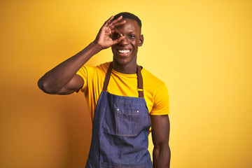 African american bartender man wearing apron standing over isolated yellow background doing ok gesture with hand smiling, eye looking through fingers with happy face.