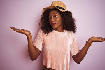 Young african american woman wearing t-shirt and hat over isolated pink background clueless and...
