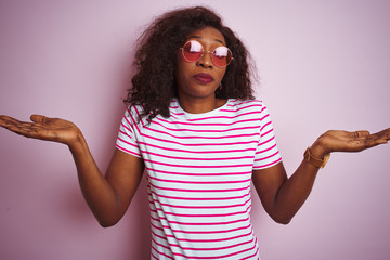 Young african american woman wearing t-shirt and sunglasses over isolated pink background clueless...