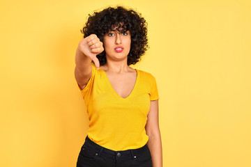 Young arab woman with curly hair wearing t-shirt standing over isolated yellow background looking unhappy and angry showing rejection and negative with thumbs down gesture. Bad expression.
