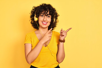 Arab woman with curly hair listening to music using headphones over isolated yellow background smiling and looking at the camera pointing with two hands and fingers to the side.