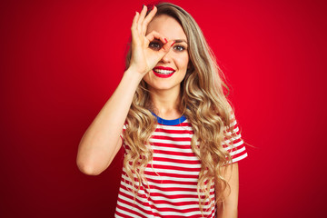 Young beautiful woman wearing stripes t-shirt standing over red isolated background doing ok gesture with hand smiling, eye looking through fingers with happy face.