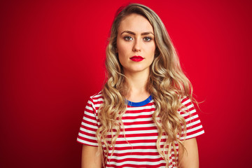 Young beautiful woman wearing stripes t-shirt standing over red isolated background with serious expression on face. Simple and natural looking at the camera.