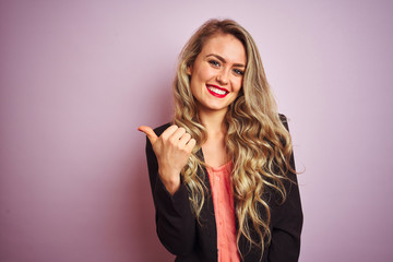 Young beautiful business woman wearing elegant jacket standing over pink isolated background smiling with happy face looking and pointing to the side with thumb up.