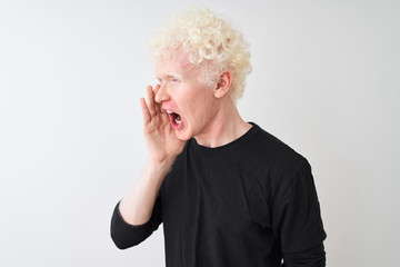 Young albino blond man wearing black t-shirt standing over isolated white background shouting and screaming loud to side with hand on mouth. Communication concept.