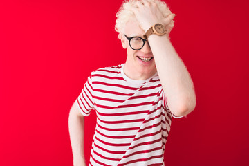 Young albino blond man wearing striped t-shirt and glasses over isolated red background surprised...