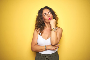 Middle age senior woman showing wrist watch over yellow isolated background serious face thinking about question, very confused idea