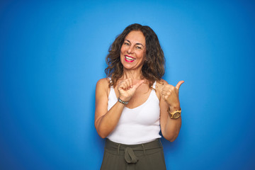 Middle age senior woman with curly hair standing over blue isolated background Pointing to the back behind with hand and thumbs up, smiling confident