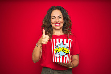 Middle age senior woman eating popcorn snack over red isolated background happy with big smile doing ok sign, thumb up with fingers, excellent sign