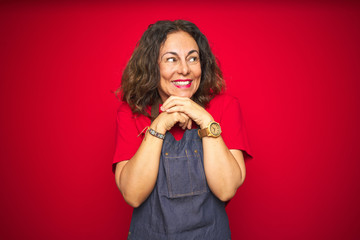 Middle age senior woman wearing apron uniform over red isolated background laughing nervous and excited with hands on chin looking to the side