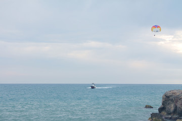 A skydiver flies with a colorful parachute tied to a speedboat on the sea against the backdrop of mountains with a blue sky. The concept of summer holidays, vacation, tourism.