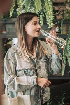 young girl drinking water from a glass bottle with an eco-bag on the background of growing pots