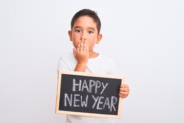 Beautiful kid boy holding blackboard standing over isolated white background cover mouth with hand...