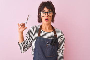 Hairdresser woman wearing apron and glasses holding scissors over isolated pink background scared...