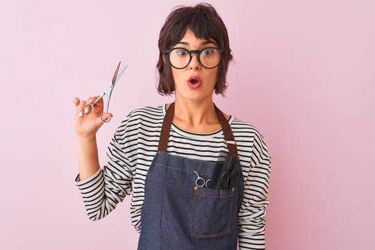Hairdresser woman wearing apron and glasses holding scissors over isolated pink background scared in shock with a surprise face, afraid and excited with fear expression